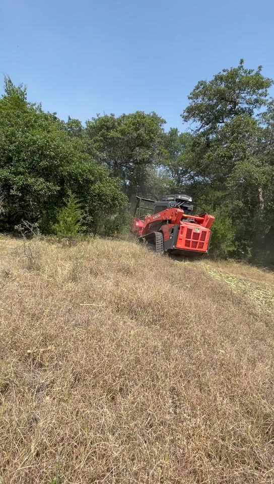 A red forestry mulcher machine on a hillside with dry grass and green trees in the background under a hazy sky.