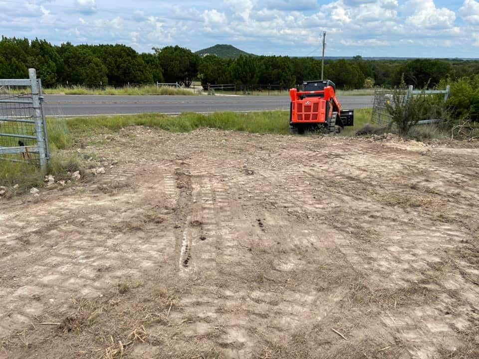 "Rural landscape with open gate, compacted dirt road, red skid steer loader on the side, green trees, and hills in the distance under a cloudy sky.